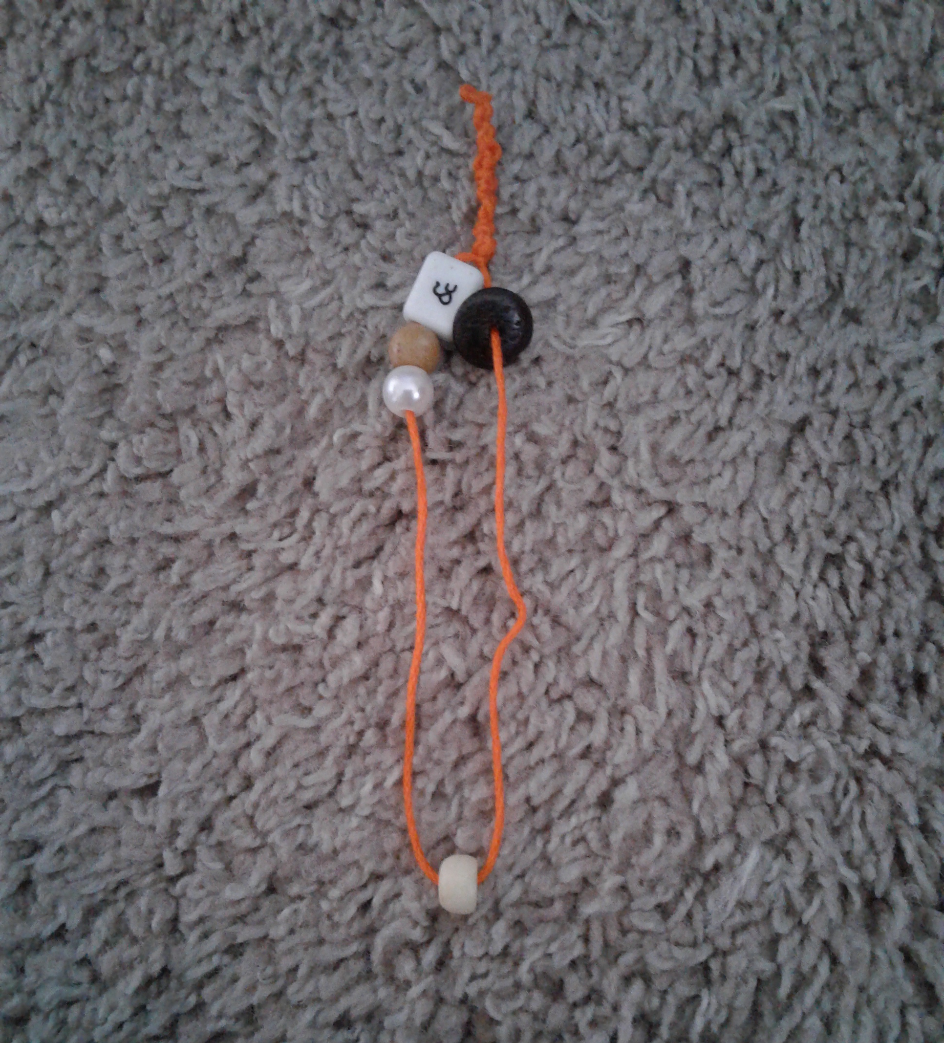 An orange bracelet with five beads: a black bead, and cream bead, a shiny bead, a brown bead, and a white bead with an ampersand on it.