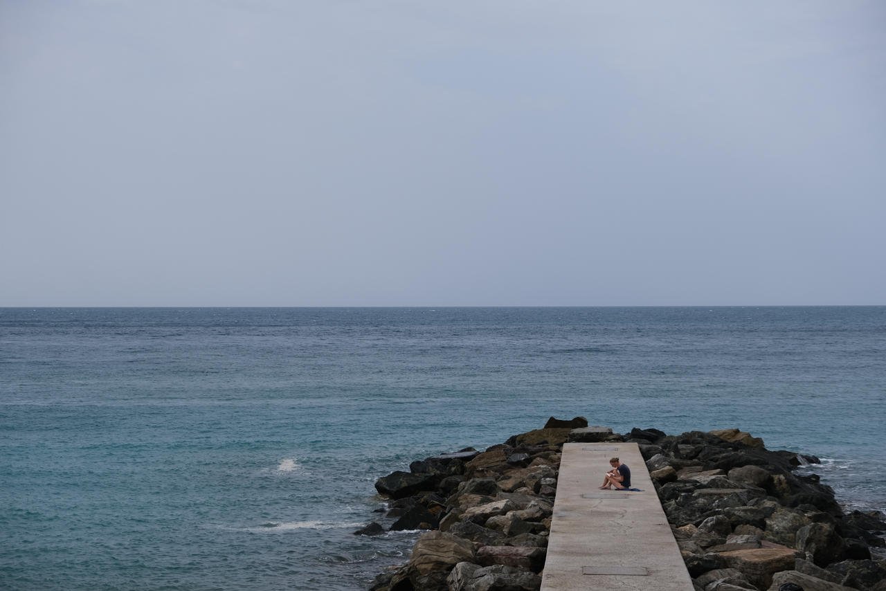 A photo of a seascape, with a rock pier on the left and a person sitting and drawing.